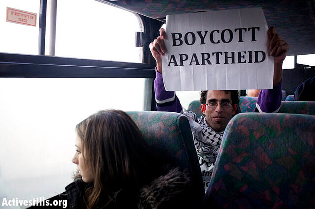 A protestor holds up a sign reading 'Boycott Apartheid' on a bus in the West Bank. (Photo: Oren Ziv/ Activestills