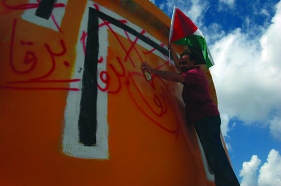 Palestinian from Burqa paints over Hebrew letters that read "Homesh First" on an evacuated settlement water tower. (Photo: Allison Deger)