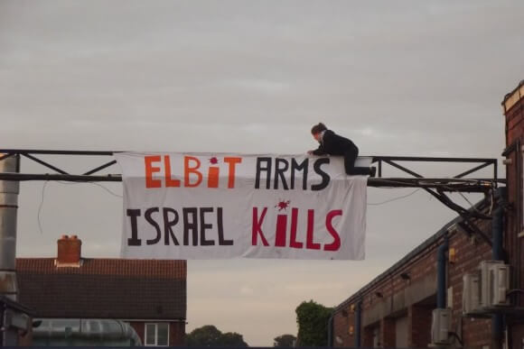 Protester hangs "Elbit Arms, Israel Kills" at shutdown Elbit factory in Birmingham. The factory produces drone engines which are exported to Israel and used over Gaza. (Photo: London Palestine Action)
