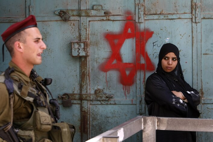 An Israeli soldier keeps guard near a Palestinian woman standing next to Star of David graffiti sprayed by Israeli settlers at an army checkpoint in the center of Hebron, May 18, 2009. (Photo: MENAHEM KAHANA/AFP/Getty Images)