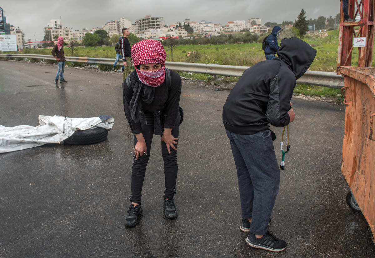 A female Palestinian protester takes coverage behind a waste container.