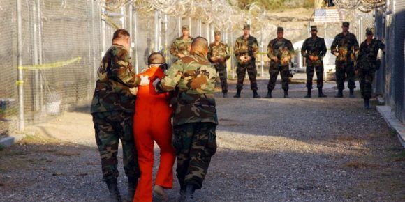 U.S. Army Military Police escort a detainee to his cell January 11, 2001 in Camp X-Ray at Naval Base Guantanamo Bay, Cuba, during in-processing to the temporary detention. The detainees will be given a basic physical exam by a doctor, to include a chest x-ray and blood samples drawn to assess their health, the military said. The U.S. Department of Defense released the photo January 18, 2002. (Photo: Petty Officer 1st class Shane T. McCoy/U.S. Navy)