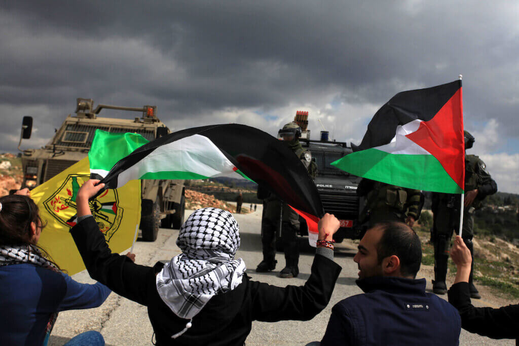 Palestinian protestors from the West Bank village of Nabi Saleh wave flags as they sit in the road in front of Israeli security forces during a demonstration against Jewish settlers near the Jewish settlement of Halamish on March 14, 2014. (Photo: Issam Rimawi/APA Images)