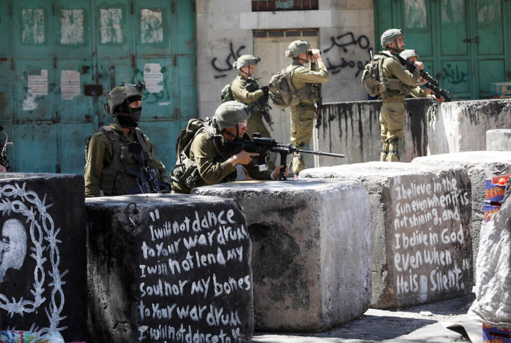 Israeli soldiers hiding behind cement blocks and taking aim with their rifles at Palestinian youth who are confronting the soldiers in Hebron after the announcement of the death of Khader Adnan in Israeli custody while he was on hunger strike.