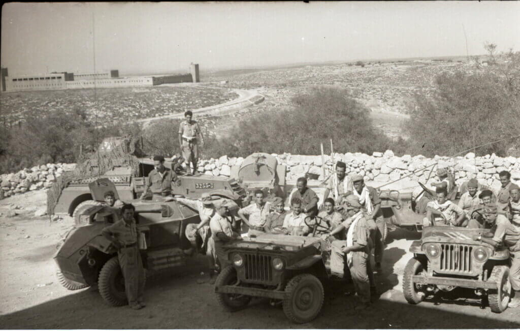 Historical photo from 1948 portraying the occupation of Iraq Suwaydan. Israeli soldiers are shown carrying rifles and riding their military jeeps as they pose for the camera, while in the background there is a stone fence and the plains of Iraq Suwaydan extending in the background, and the old British Police station can be seen in the distance.