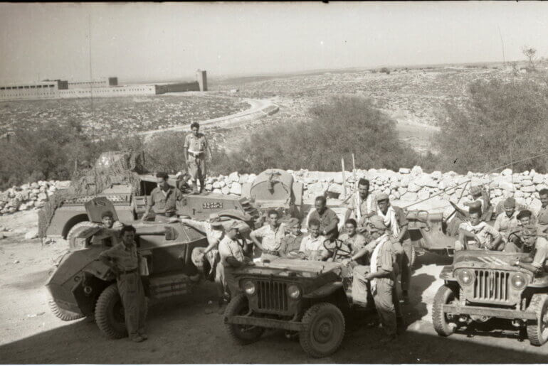 Historical photo from 1948 portraying the occupation of Iraq Suwaydan. Israeli soldiers are shown carrying rifles and riding their military jeeps as they pose for the camera, while in the background there is a stone fence and the plains of Iraq Suwaydan extending in the background, and the old British Police station can be seen in the distance.