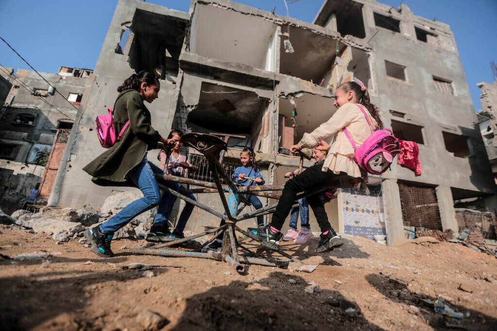 Young girls in Gaza play on a the broken remains of a carousel after the bombing of their home in Gaza, with the dilapidated bombed-out remains of their home in the backdrop.