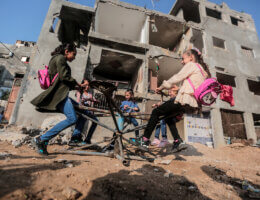 Young girls in Gaza play on a the broken remains of a carousel after the bombing of their home in Gaza, with the dilapidated bombed-out remains of their home in the backdrop.