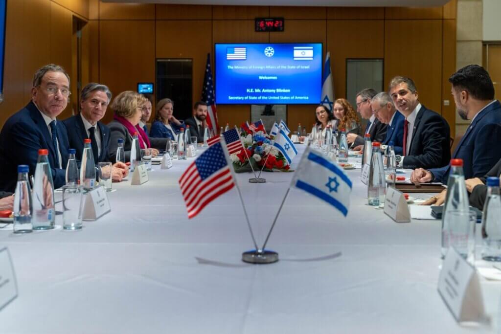Secretary of State Antony Blinken and Israeli Foreign Minister Eli Cohen sit across from each other at a large rectangular table. People of their delegations are around them. On the table stand little U.S. and Israeli flags. Behind them is a blue screen that reads "The Ministry of Foreign Affairs of the state of Israel welcomes H.E. Mr. Antony Blinken, Secretary of State of the United States of America".