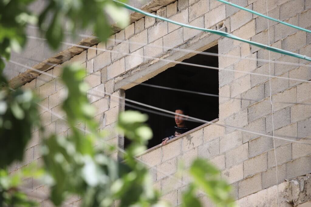 A young man looks outside of the window of an unfinished building in the Aida refugee camp, with electric wires in the foreground. (Malik Hamamra/Mondoweiss) Aida Refugee Camp, occupied West Bank, May 2023.
