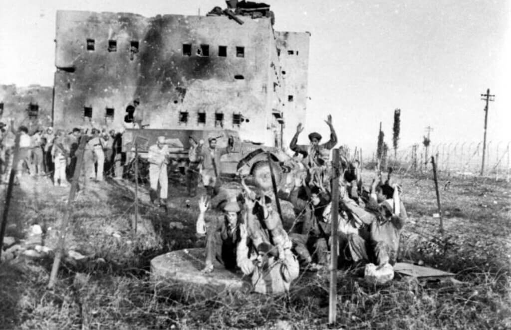 Historical photo of the Nakba shows Israeli soldiers pointing guns at prisoners, who are gathered together with hands raised above their heads in surrender. In the background the burnt building of the British police station of Iraq Suwaydan can be seen.