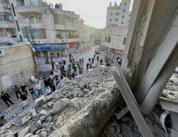 A photo take from within a blown-out remains of an apartment in Ramallah detonated by the Israeli army, pointing towards the outside where a wall used to be. A crowd of people gathering below to witness the destruction can be seen.