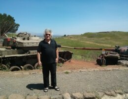 Ephraim Laor standing in the occupied Syrian Golan Heights in April 2014, with two tanks behind him and rolling green hills in the background.