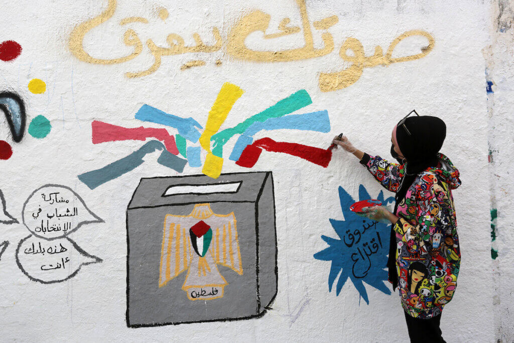 A girl in Gaza paints a mural depicting a ballot box with an eagle symbol painted on it, with a Palestinian flag emblazoned on the eagle's chest and the word "Palestine" written at the eagle's feet, and with multi-colored hands slipping ballots into the box.