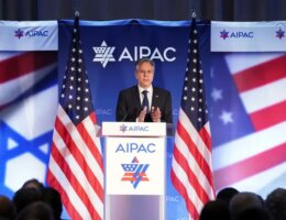 An image of US Secretary of State Antony Blinken standing on a podium at AIPAC, flanked by two American flags and with the AIPAC logo behind him.