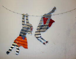 Corrugates steel plates shaped into images of children playing and dangling from barbed wire, and painted over.