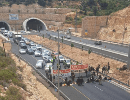 A photo from a distance of activists shutting down a main highway connecting West Bank settlements to Jerusalem. The activists are seen sitting in one of the highway lanes with a blocking a line of cars, with two large unfurled banners. One banner reads, "Apartheid All Exists," while the other banner is in Arabic, and reads "An obstacle in the face of fascism."