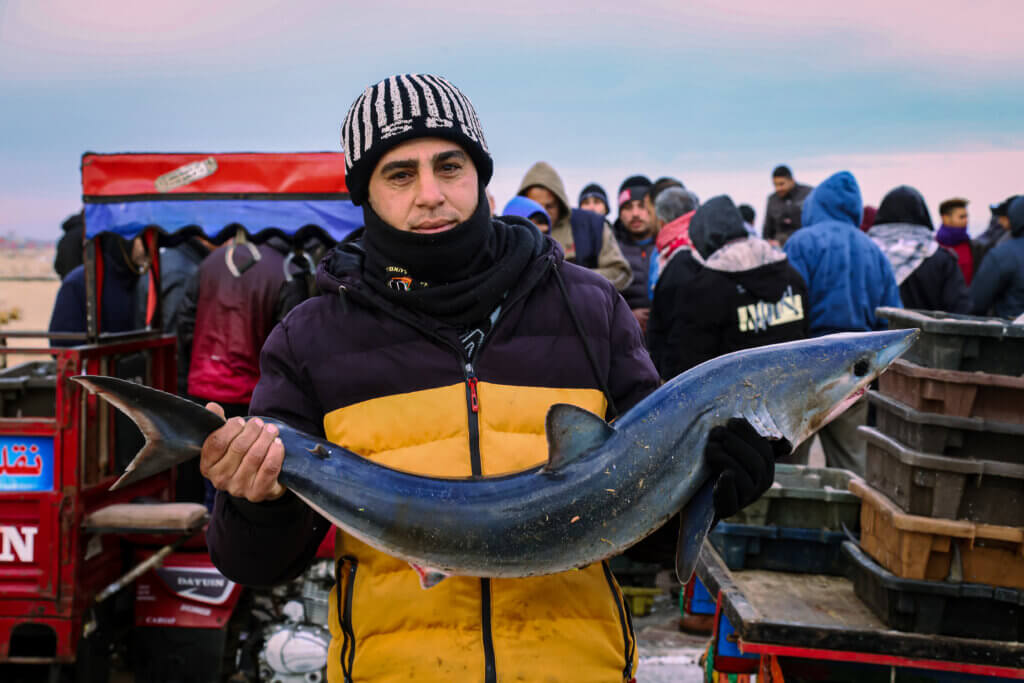 A fishmonger in Gaza holds up a large fish in both hands with Gaza's seaport in the bakcground.