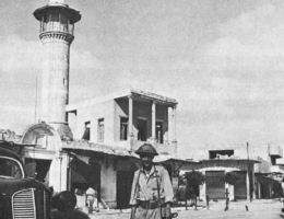A balck-and-white photo of an Israeli soldier posing in front of the Dahmash Mosque in Lydd in 1948, standing next to a civilian vehicle and carrying a machine gun.