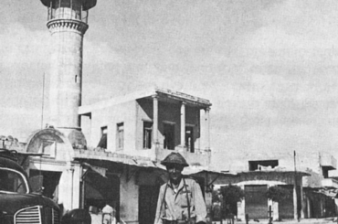 A balck-and-white photo of an Israeli soldier posing in front of the Dahmash Mosque in Lydd in 1948, standing next to a civilian vehicle and carrying a machine gun.
