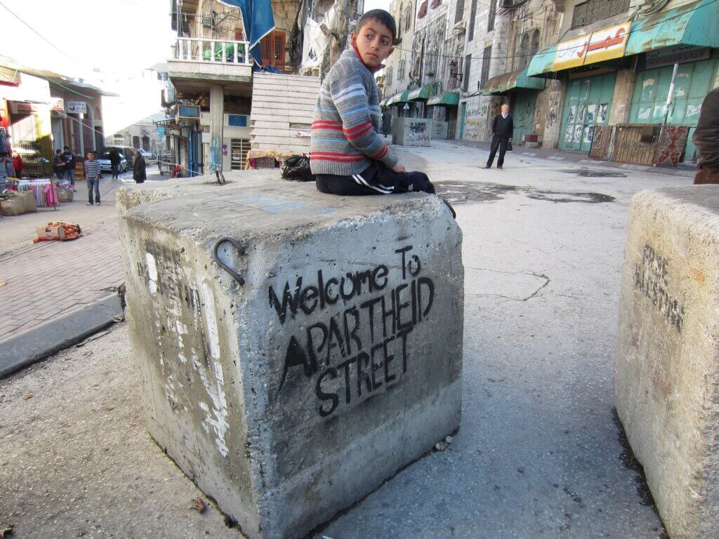 A Palestinian child sits on a barrier blocking a street in the middle of the West Bank city of Hebron that Palestinians are prevented from using while illegal Israeli settlers have free movement under protection of the military. (Photo: Wikimedia/Austin 202)