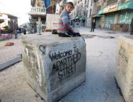 A Palestinian child sits on a barrier blocking a street in the middle of the West Bank city of Hebron that Palestinians are prevented from using while illegal Israeli settlers have free movement under protection of the military. (Photo: Wikimedia/Austin 202)
