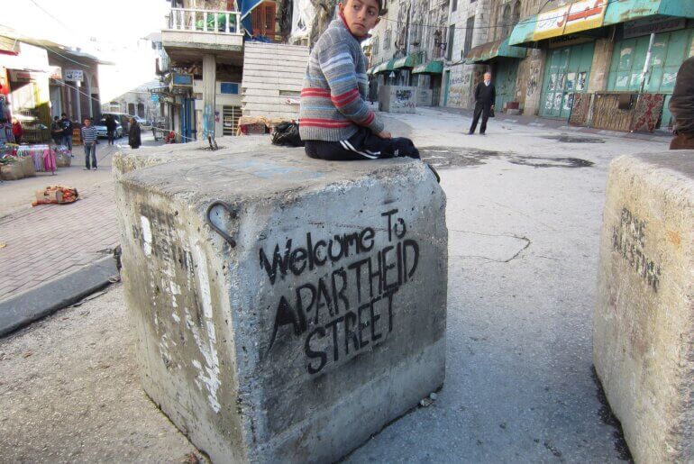 A Palestinian child sits on a barrier blocking a street in the middle of the West Bank city of Hebron that Palestinians are prevented from using while illegal Israeli settlers have free movement under protection of the military. (Photo: Wikimedia/Austin 202)