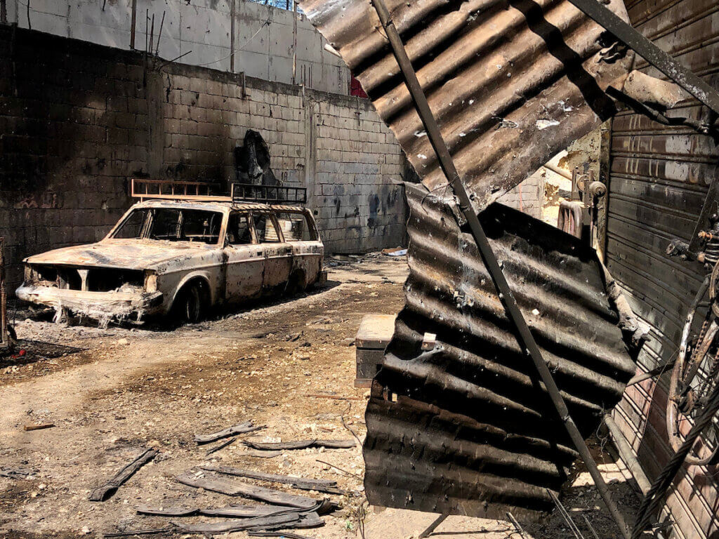 Scenes of destruction, including a dilapidated and burned car that sits in the middle of a narrow alleyway in Ein al-Helweh refugee camp, with bent and rusted corrugated steel plates in the foreground.