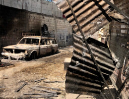 Scenes of destruction, including a dilapidated and burned car that sits in the middle of a narrow alleyway in Ein al-Helweh refugee camp, with bent and rusted corrugated steel plates in the foreground.