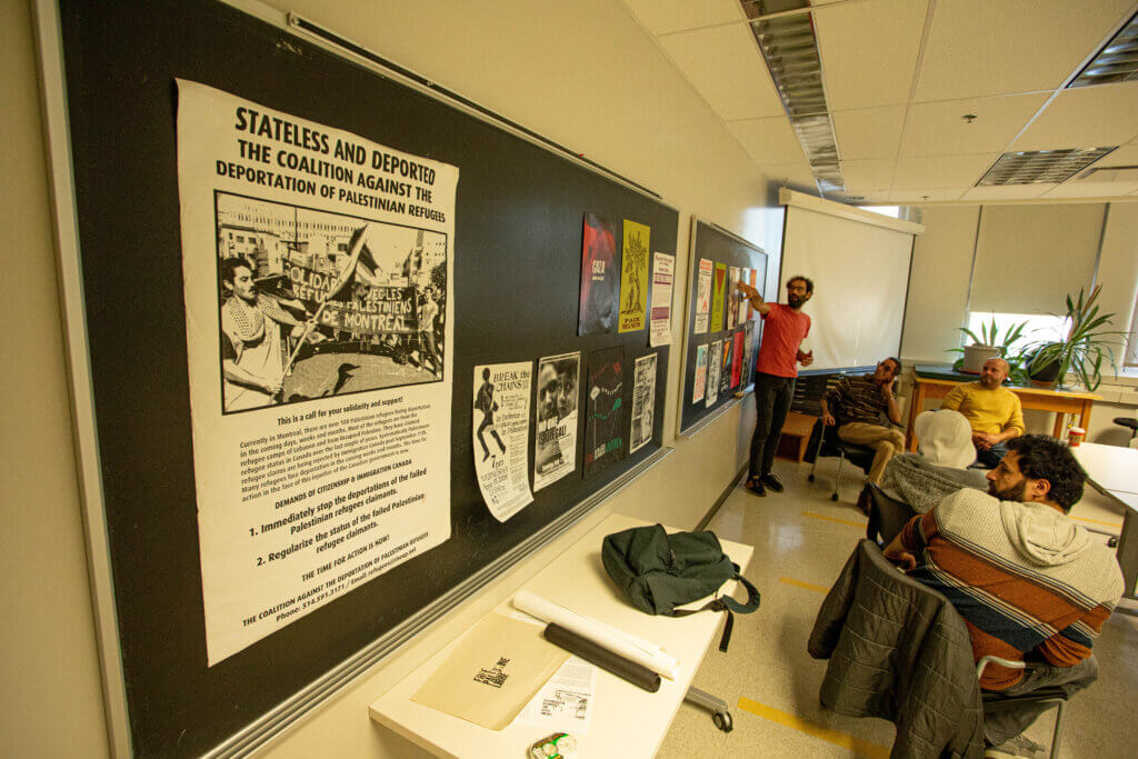Conference at Concordia to mobilize support for Youssef and other non-status Palestinians in the context of the broader #StatusForAll movement. The picture depicts an activist in a salmon-colored t-shirt giving a presentation and pointing to posters on a wall. One of the posters reads: "Stateless and deported: the coalition against the deportation of Palestinian refugees."