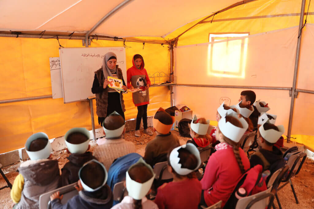 Students sit in rows sporting white caps as two teachers stand before them during class session, which is being held under a tent over the ruins of a demolished school in Masafer Yatta.