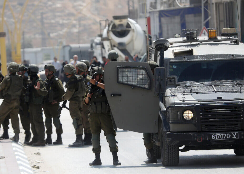 Israeli soldiers gather around a roadblock, while one soldier perches behind an army jeep and takes aim towards the camera with his rifle.