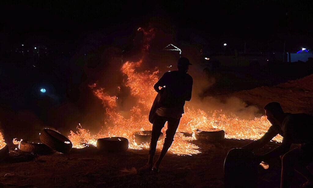 A nighttime photo showing the sillhouette of a masked demonstrator holding a car tire while other tires are burning in the background and flames light up the backdrop.