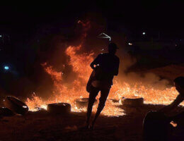 A nighttime photo showing the sillhouette of a masked demonstrator holding a car tire while other tires are burning in the background and flames light up the backdrop.