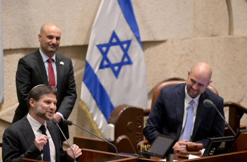 Finance Minister Bezalel Smotrich speaking at a podium in the Knesset, with the Israeli flag behind him.