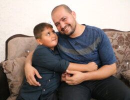 Tamer Za'anin (right) hugging his son Hassan (left) as they sit on a couch and smile at the camera. Hassan was conceived through Tamer's smuggled sperm while he was in Israeli prison.