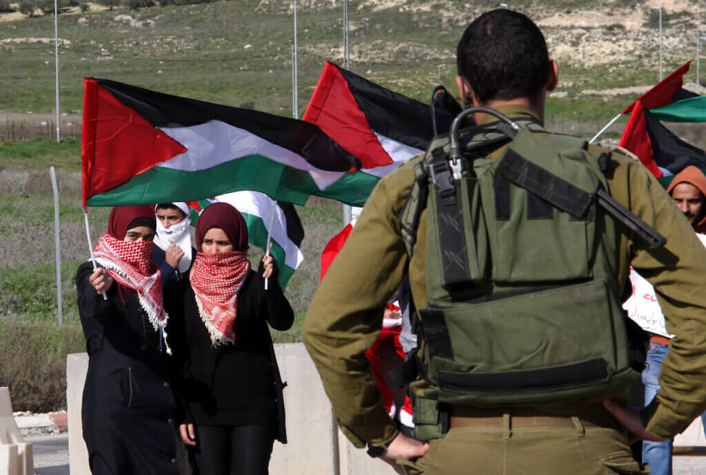 An Israeli soldier stands in front of Palestinian women waving Palestinian flags during a demonstration at the Huwwara checkpoint near Nablus in the West Bank, January 1, 2015. (Photo: Nedal Eshtayah/APA Images)
