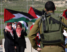 An Israeli soldier stands in front of Palestinian women waving Palestinian flags during a demonstration at the Huwwara checkpoint near Nablus in the West Bank, January 1, 2015. (Photo: Nedal Eshtayah/APA Images)