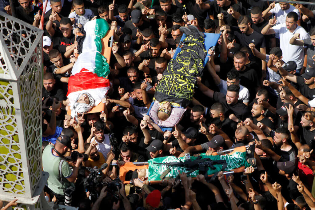 Mourners carry the bodies of three Palestinian resistance fighters, Rafat Khamayseh, Mahmoud al-Sa'di, and Mahmoud Ararawi, killed during an Israeli raid on Jenin refugee camp, September 20, 2023. The photo is an overhead shot of the crowd of mourners carrying the three bodies ons tretchers. One is draped in a Palestinian flag, the other in a black and yellow Islamic Jihad flag, and the third in a green Hamas flag.