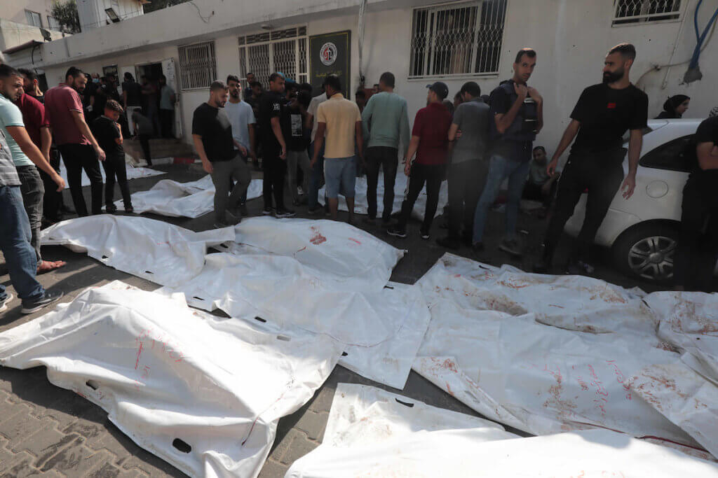 People stand by the bodies of victims of Israeli air strikes outside the morgue of al-Shifa hospital in Gaza City on October 12, 2023. The victims' bodies are lined on the ground and covered with white tarps.