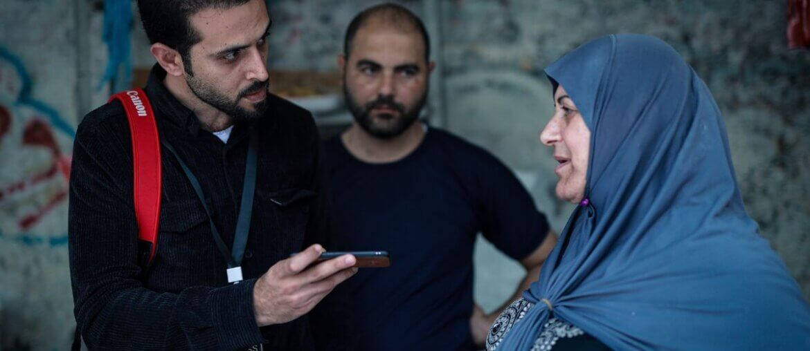 Tareq Hajjaj (left) during one of his field visits reporting for Mondoweiss. He is recording an interviewee on his phone with a camera slung over his shoulder as he interviews a woman in Gaza.