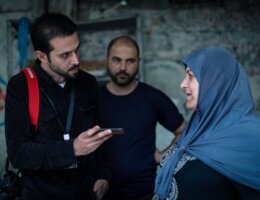Tareq Hajjaj (left) during one of his field visits reporting for Mondoweiss. He is recording an interviewee on his phone with a camera slung over his shoulder as he interviews a woman in Gaza.