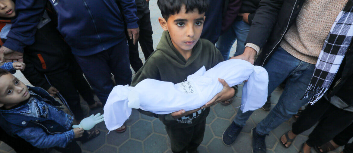 A young Palestinian boy holds the shrouded body of a Palestinian infant who was killed by Israeli airstrikes in Gaza in Deir al-Balah in the central Gaza Strip. (APA Images)