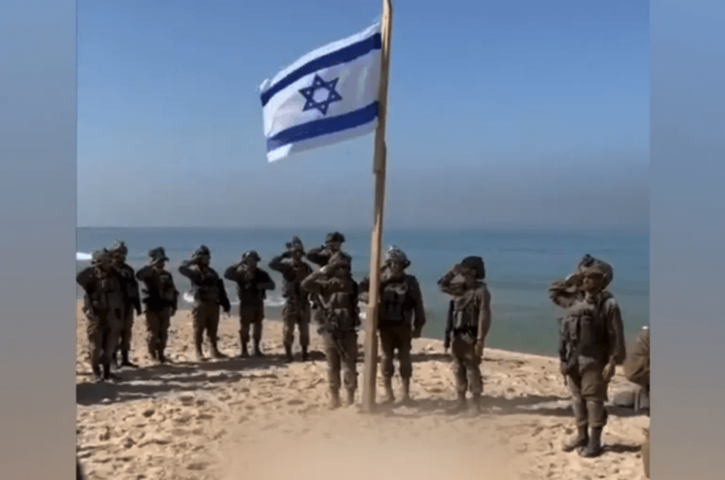 Screenshot from a video showing Israeli soldiers saluting the Israeli flag while singing the Israeli national anthem on a beach in Gaza in November 2023