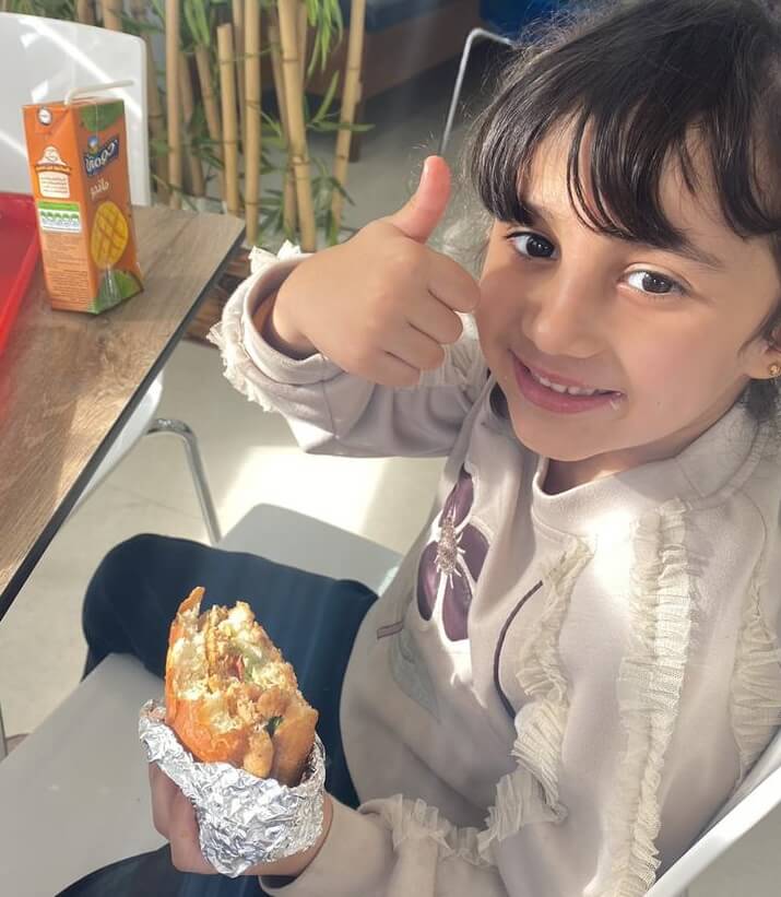 A young child eating a sandwich with a smile on their face giving a thumbs up.