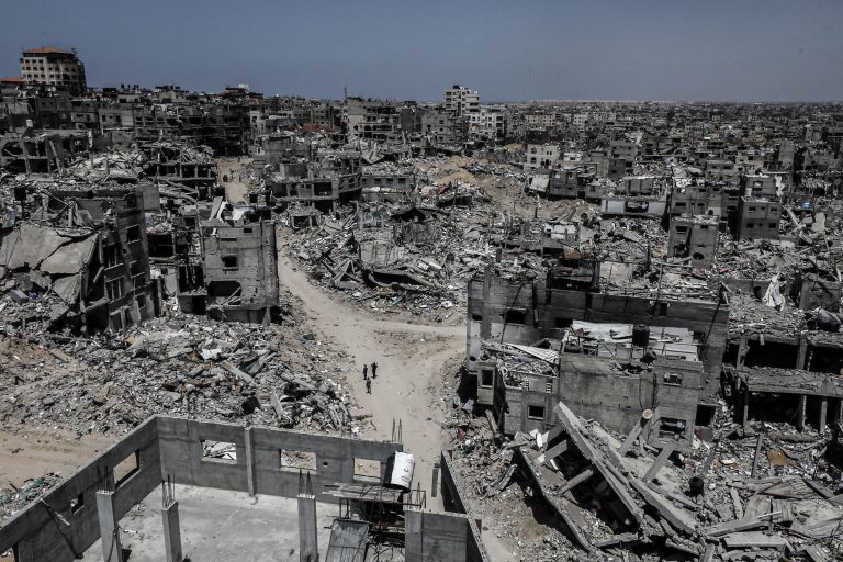 도시를 조감하듯 찍은 사진. 대부분의 건물이 반파 혹은 완파되어 있다. 비포장 상태로 보이는 도로에 세 사람이 서 있는 모습이 작게 보인다.