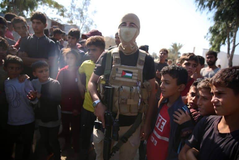 Palestinian militants of the Izz al-Din al-Qassam Brigades, the military wing of Hamas, stand guard next to a crowd watching the transfer of released Israeli hostages to the Red Cross in the south of Deir el-Balah in the central Gaza Strip, October 13, 2025. (Photo: Omar Ashtawy/APA Images)