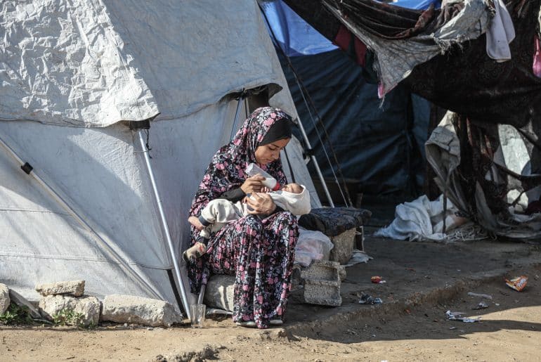 A Palestinian woman bottle feeds a baby outside a tent in a displacement camp in Gaza