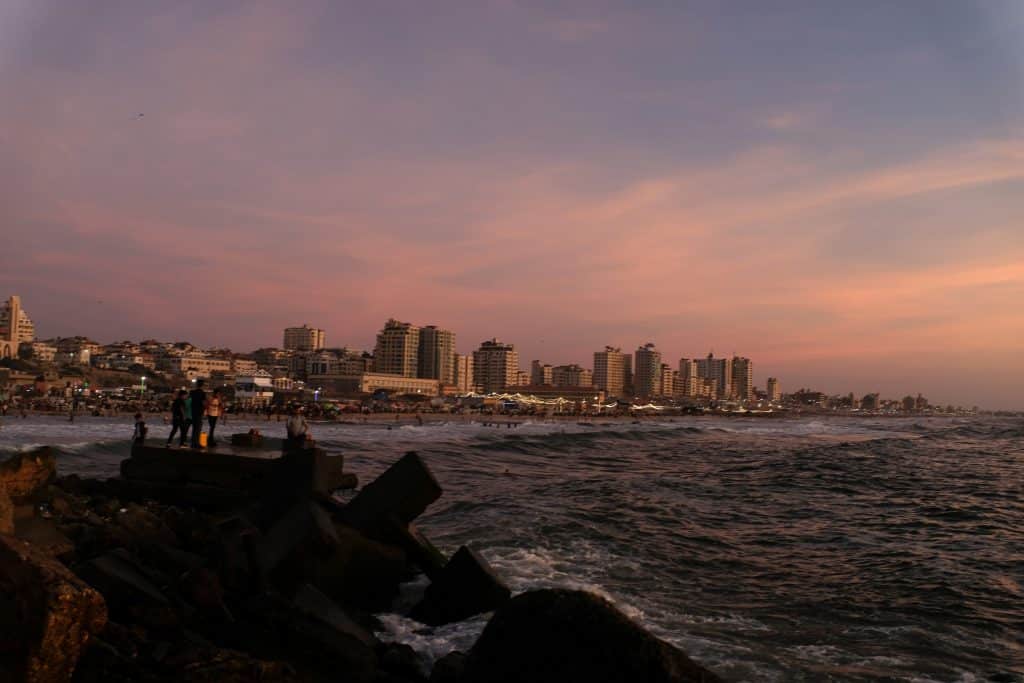 People gather along the rocky edge of Gaza’s coast as the city skyline fades into dusk, July 11, 2022. (Photo: Mahmoud Nasser)
