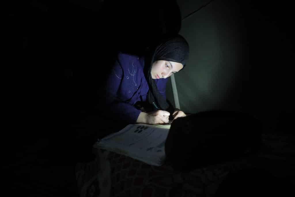 Dana Mohammed Abu Dalfa, a Palestinian high school student, reviews her lessons using the light of a mobile phone inside a displacement tent in the Gaza Strip, June 20, 2026. (Photo: Hassan Jedi /APA Images)